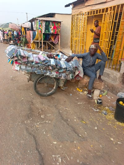 A young man sits beside a cart overflowing with packaged phone cases. He wears a gray outfit and sandals, resting his hand on his head with a relaxed posture. Behind him, a man stands in a building with yellow bars, using a cell phone. A makeshift structure displays colorful clothes nearby. The ground is littered with debris, including what appears to be corn kernels and fruit peels. A black bucket filled with yellow items sits in the lower right corner.