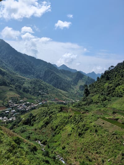 This is a high-angle, long shot photograph of a verdant, mountainous landscape in Minh Son Commune, Vietnam. The scene is dominated by lush green mountainsides covered in dense foliage and trees, with terraced fields carved into the slopes. In the valley below, a small village with numerous houses with dark roofs is nestled amidst the greenery. A winding road is visible within the village. A small stream flows through the foreground, flanked by dense vegetation. The sky above is a clear blue with scattered white clouds, suggesting a daytime setting. The overall impression is one of natural beauty and rural life in a mountainous region.