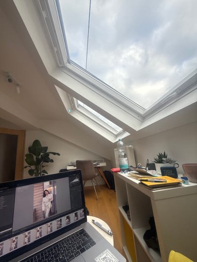 Here is a description of the image:

The image shows a view of a room in a building with a slanted roof and a skylight, letting in daylight and view of a cloudy sky. The room contains a desk or shelf unit on the right, and a laptop computer is placed on it. The screen of the laptop shows an image editing software with an image of a person in white clothing. Near the computer there is a pen on the table. A plant stands on the floor.
The image appears to be taken in an indoor setting, possibly in Ampfing, Germany, given the OCR of the image.