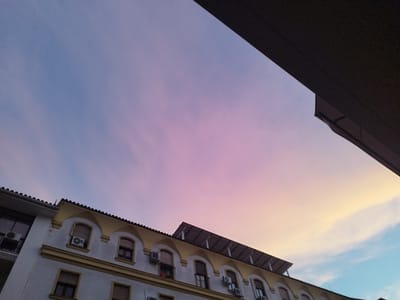 A low-angle shot captures the sky over buildings in Jerez, Spain, during what appears to be sunset or dawn. The sky transitions from a pale blue at the top right to a soft pink and then a pale yellow towards the horizon. Wispy clouds are visible. In the foreground, the upper floors of two buildings are visible. The building on the left has a gray tiled roof and a white facade with several windows and what appears to be an air conditioning unit. The building to the right has a yellow and white facade with a tiled roof. This building features arched windows, some with air conditioning units, and a set of solar panels installed on its roof. A dark, angular structure, possibly an overhang or another building, occupies the upper right portion of the frame.