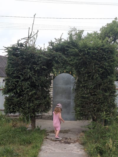 In Perliavka, Ukraine, a young child wearing a pink jumpsuit and a pink cap walks away from the camera on a cracked concrete path. The child is framed by two large, manicured evergreen trees that arch over the path, creating a natural gateway. Behind the trees, a large, weathered metal gate stands in front of a light-colored building. The scene is set outdoors with green grass and foliage growing along the sides of the path. Overhead, power lines are visible against a cloudy sky.
