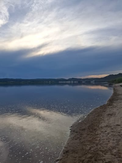 The image depicts a serene natural landscape at what appears to be the edge of a body of water, possibly a river or lake, adjacent to a sandy shore. The sky is overcast with dramatic, dark clouds interspersed with areas of lighter, wispy clouds that reflect sunlight. The water surface is calm and dark blue, with a scattering of small white foam or bubbles visible near the shore and reflecting the sky. The far bank of the water is lined with a dark, undulating treeline, with some lighter-colored formations or bluffs visible in the distance. The foreground consists of a sandy beach with tire tracks pressed into the sand, suggesting recent human activity. The overall mood is tranquil and somewhat moody due to the overcast sky. The scene is located in the vicinity of Малая Сазанка, Russia.