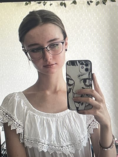 A young woman with light brown hair pulled back and wearing glasses is taking a selfie in a room. She is wearing a white off-the-shoulder top with lace trim. She is holding a smartphone with a black and white graphic design of a face on the case. The background appears to be a white wall with a textured pattern, and there are some green leaves hanging from the top of the frame. The image does not contain enough information to determine the location in the city of Pruszcz Gdański, Poland.
