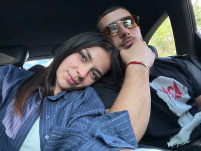 A selfie shows a couple in a car. The woman has long dark hair and wears a blue and white striped shirt. The man wears sunglasses and a black t-shirt with a white and red graphic. The car is black. Through the windshield, trees and the sky are visible. The location, Хаджийско, Bulgaria, is not discernible from the image.