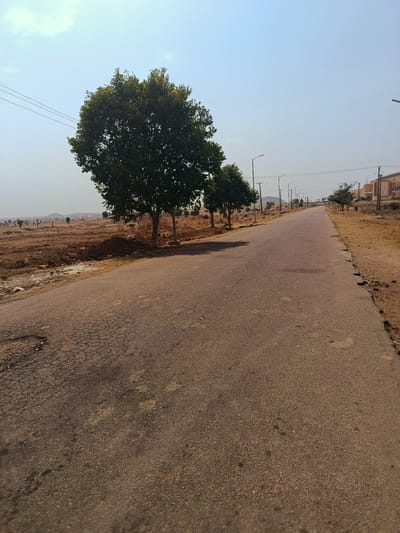 The image depicts a straight, asphalt road stretching into the distance. The road surface is worn and cracked. On either side of the road, the land is barren with dry, brown soil. Sparse vegetation and scattered trees line the road. Streetlights are visible along the road's edge. In the distance, low hills can be seen. The sky is clear and pale blue. Overhead, utility lines are strung between poles. The scene is located in Rafin Pa, Nigeria. No people are visible.