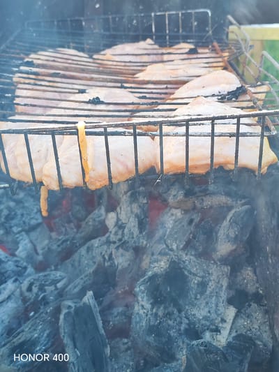 The image shows chicken breasts cooking on a grill over hot coals. The grill is made of metal bars, and the chicken pieces are arranged in rows on the upper rack. Some of the chicken pieces have a slightly orange hue, possibly from a marinade. Smoke is visible rising from the grill. Below the grill, glowing red coals are visible through the grates, indicating intense heat. The overall setting appears to be outdoors, with a hint of greenery in the background on the right side. The bottom left corner of the image displays the text "HONOR 400". There are no people visible in the frame. This scene is likely taking place in a residential backyard or a picnic area. There is no specific information in the image to pinpoint the location within the city of Эрхирик, Russia.
