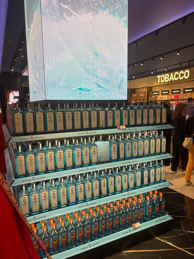 The image shows a display of Bombay Sapphire gin bottles in a duty-free shop, likely at an airport. The location is identifiable as Ferno, Italy due to the "CASSA TICA" checkout sign. Several shelves are stacked with bottles of Bombay Sapphire, each with a blue label. In the background, a "TOBACCO" sign is visible. Some people are present, observed from the back, indicating that the scene is populated. A large digital screen is positioned to the left, showing a light-colored abstract image. There is a red fabric or decorative element in the lower right corner.