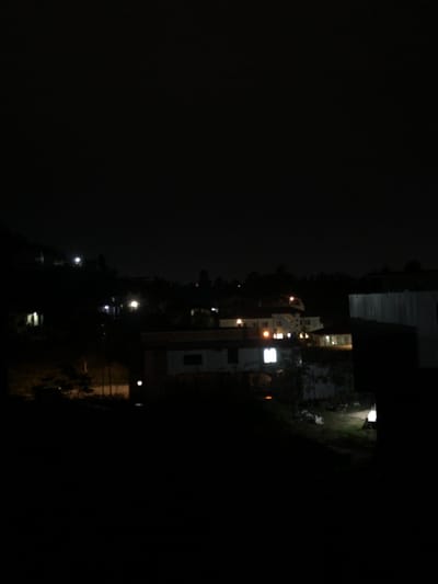 A night scene presents several structures with illuminated windows and outdoor lights. Some appear to be houses or buildings on a hill, descending into darker foliage. The left side of the image is predominantly dark, suggesting a vast expanse of night sky or obscured land. Streetlights are visible along a road or path, further defining the residential or developed area.