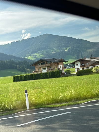 The image shows a rural mountainous landscape in Reitdorf, Austria, likely taken from a vehicle. The scene is dominated by a steep, forested mountain on the left, partially obscured by a bright, cloudy sky. In the mid-ground, there are several multi-story buildings with traditional European architecture, featuring wooden balconies and white or beige facades. These buildings are set against a backdrop of rolling green hills and more dense forest. A well-maintained, vibrant green lawn runs along the right side of the image, adjacent to a paved road. The road has white lane markings, indicating it is a public thoroughfare. A white cylindrical object, possibly a reflective marker or bollard, stands on the edge of the lawn next to the road. The sunlight suggests it is daytime, and the clear sky with scattered clouds indicates fair weather. The overall impression is one of a peaceful, scenic alpine village or resort. No people are visible in the frame, and there is no visible text.