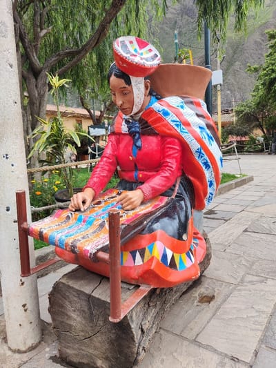 A statue depicts a woman with a red jacket and a patterned shawl, seated at a colorful loom. She wears a white head covering and has a red and white hat perched on her head. The loom is supported by a log bench. The scene is set outdoors on a paved area, with lush greenery, trees, and a mountain visible in the background. A concrete post is positioned to the left of the statue.