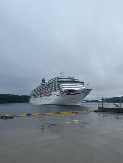 A large white cruise ship, identified as the Arcadia by the text on its hull, is docked at a pier. The ship has a prominent Union Jack flag design on its bow and is emitting a plume of smoke from a stack near its stern. The sky is overcast and gray. The pier in the foreground is wet and appears to be made of concrete. Yellow and blue bollards are visible along the edge of the pier. In the background, to the left of the ship, is a green, tree-covered hillside. To the right of the ship, beyond a chain-link fence, there are some industrial structures and a hilly landscape. A red and white life preserver hangs on the fence. The scene suggests the ship is at a port in a coastal area under a cloudy sky. The presence of the Arcadia cruise ship in Bodø, Norway, indicates the location is likely the port of Bodø.