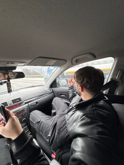 This image captures the interior of a vehicle, specifically the front cabin, in Gabarnitsa, Bulgaria.

A male individual is seated in the front passenger seat, observed from a rear-left perspective. He has short, dark brown hair and a beard, and is wearing a black puffer jacket and dark grey or black jeans. His legs are propped up onto the dashboard. His right arm is raised, with his hand extended and the middle finger prominently displayed towards the vehicle's right side window.

In the foreground, the left hand of another individual, presumably the driver or photographer, is visible, holding a black-cased smartphone. The driver's face is partially reflected in the rearview mirror, along with a dangling car air freshener.

The car's dashboard is dark grey, featuring a radio unit with a red digital display showing "0223" and various buttons and controls. An air vent is visible on the passenger side. A black can, possibly a beverage, rests in a cup holder near the passenger's feet. Through the windshield, a road with a white guardrail and distant vegetation is visible. Through the passenger side window, a blurry white structure with a yellow stripe is discernable. The car's interior ceiling is light grey.