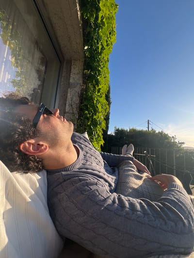 A man lies on his back on a white striped surface, possibly a mattress or a sun lounger. He is wearing sunglasses and a blue cable-knit sweater, with a patterned gray cushion resting on his chest. His legs are extended with white socks visible.
The scene appears to be outdoors in Gondarém, Portugal. A patch of green foliage overhangs a window, creating a natural edge to the scene. Beyond the foliage, a clear blue sky dominates the upper portion of the image. A utility pole with wires is also visible amongst distant trees. The sunlight is bright, causing some lens flare. The window appears to reflect the greenery.