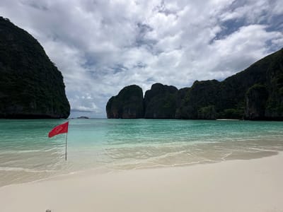 This image depicts a tropical beach scene, likely in Ao Nang, Thailand, characterized by its pristine white sand, clear turquoise waters, and dramatic limestone cliffs covered in lush vegetation. A red flag with a white symbol, possibly indicating a warning or prohibition, is planted in the shallow water near the shore. In the distance, a large white boat is visible on the calm sea, and several towering, verdant cliffs form a natural amphitheater around the bay. The sky is filled with a dramatic display of clouds, suggesting an overcast or partly cloudy day, but the overall atmosphere is serene and picturesque. The gentle waves lapping at the shore create a sense of tranquility.
