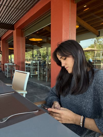 A woman with long, dark hair wearing a grey sweater is seated at a table, appearing to be using a cell phone. She has two bracelets on her left wrist. The table has a dark grey surface and a brown woven placemat with silverware. The setting appears to be an open-air restaurant with a view to the outside, possibly in Pueblo Nuevo de Jasso, Mexico, given the request. The restaurant has a structure with red-brown pillars and wooden beams. Visible through the restaurant are trees and possibly other buildings.