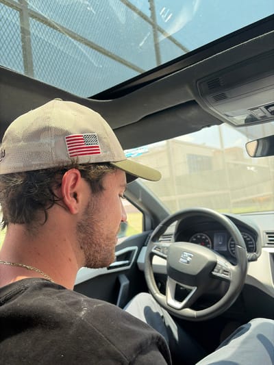 The image shows a man in the driver's seat of a car. He is wearing a light-colored baseball cap with an American flag patch, and a black shirt. A gold chain is visible around his neck. The car's steering wheel is in the foreground. The background is blurry and indistinct. The location is unknown, and there is no evidence to place it in Santa Cruz Tehuixpango, Mexico.