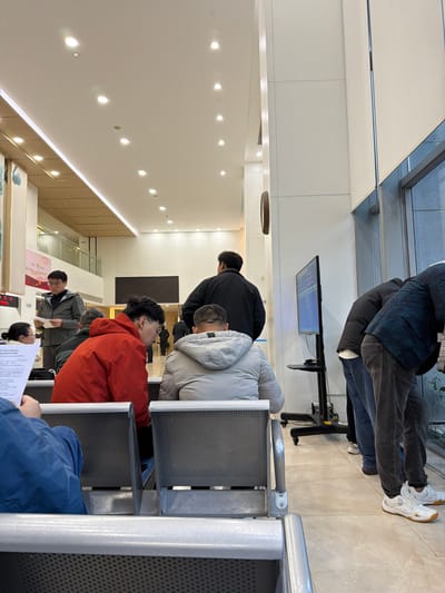 The image presents an interior view of a modern public facility located in Jianye District, China. The environment features a high white ceiling equipped with multiple recessed circular lights and a continuous linear light fixture along the upper left wall. The floor is covered with light-toned tiles. Large windows fitted with vertical blinds are visible on the right side, illuminating the space.

Approximately ten individuals are visible, predominantly adults dressed in casual winter clothing, including various padded jackets. In the foreground, a person wearing a blue jacket is partially visible, seated on a metallic waiting chair with perforated backing, holding a white document. Behind this individual, a person in a red hooded jacket, wearing glasses, sits and looks downwards, presumably at a document or device. Adjacent to them, another person in a light grey padded jacket is seated with their back to the camera.

Further to the left, a man in a dark jacket and glasses stands, examining a white document. Behind him, a woman with dark hair is partially visible, seated. In the mid-ground center, a man in a dark jacket stands facing away. To the right, near the windows, two men are positioned standing and leaning forward; one wears a dark jacket and blue jeans, the other a patterned dark top and dark jeans, both sporting white athletic shoes. They appear to be looking out or interacting with something near the window.

A large flat-screen monitor on a black mobile stand displays text and data, positioned near the windows. On the far left wall, partially visible, are posters with Chinese characters and red digital numbers, likely a queue number display. A circular, textured brown object is mounted on a white pillar in the mid-ground. The overall scene depicts a busy administrative or service center where individuals are waiting or engaged in processing paperwork.