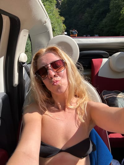 A fair-skinned woman with blonde hair and sunglasses is sitting in the passenger seat of a convertible car, taking a selfie. She is wearing a black bikini top. The car is parked on a road with trees and a hillside in the background. There are other cars and a construction vehicle visible on the road in the distance. The scene appears to be in Breitnau, Germany, as indicated by the presence of trees and the hilly terrain typical of the region. The woman is puckering her lips as she takes the photo. A Louis Vuitton bag is visible in the seat next to her.