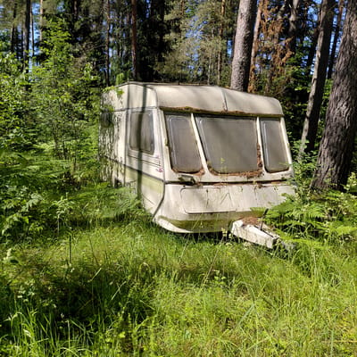 The image shows an abandoned, weathered white caravan in a dense forest. The caravan is tilted on its side, partially overgrown with grass and foliage, suggesting it has been left in this location for a significant period. The scene is set in a natural environment with tall trees and thick undergrowth. Sunlight filters through the trees, indicating it is daytime, likely a sunny day given the brightness. There are no people visible in the image. The caravan's windows are opaque or dusty, and some debris, possibly pine needles, are visible around the window frames. A light green striped decal runs along the side of the caravan, and there are some indistinct logos or emblems on the side. The overall impression is one of decay and neglect within a natural, overgrown setting. The location context provided is Skuķīši, Latvia.