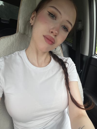 A young woman with fair skin, dark hair braided to her right, and light brown eyes is pictured inside a vehicle. She is wearing a white t-shirt. The woman has winged eyeliner and pink blush on her cheeks. Her hair is tied with a dark elastic band. The background shows the interior of a car, including a fuzzy white seat cover on the left and black leather on the right, with a black seatbelt visible. The scene is outdoors, as indicated by the greenery visible through the car window on the right. The lighting suggests it is daytime. The image was taken in Gavrilovka, Russia.