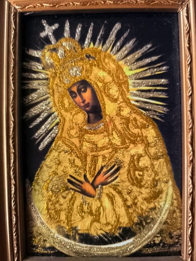 This is a framed religious icon depicting the Virgin Mary. She is shown from the chest up, with her head tilted down and her hands crossed over her chest. She wears a golden crown and a richly adorned golden robe, accented with glitter. A radiant halo surrounds her head, also embellished with glitter. The background is a dark, deep blue. The frame is ornate and metallic, appearing to be gold-toned. The overall impression is one of reverence and divine beauty. The image does not provide any specific cues about the time of day, weather, or geographic location, beyond its context in Almere, Netherlands. No text is visible in the image.