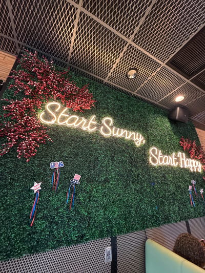 The image depicts a wall adorned with a green, artificial hedge, punctuated by clusters of red decorative berries on branches. Two lines of white neon text are prominently displayed: "Start Sunny" and "Start Happy." Below the neon text, several small, patriotic-themed decorations are attached to the hedge, including a star with the word "Proud" and ribbons in red, white, and blue, as well as a miniature Uncle Sam hat with similar ribbon accents. A person with curly brown hair is partially visible in the lower right corner of the frame, seated on a light green upholstered bench. The ceiling features a metallic grid structure with recessed lighting fixtures. The overall ambiance suggests a restaurant or cafe setting, likely in Naples, Florida, USA, given the patriotic decorations, possibly in celebration of an American holiday.