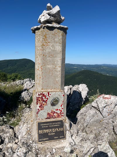 The image depicts the summit of Veliki Vukan, a mountain located in Petrovac-na-Mlavi Municipality, Serbia. A stone monument, topped with a stack of rocks, stands prominently in the foreground. The monument is adorned with a mosaic of red and white tiles and a circular reflective surface. Below the mosaic, a metal plaque displays text in Cyrillic script, identifying the location as "Veliki Vukan" and stating its elevation as "825M". The plaque also includes dates, "50 ГОДИНА" and "1960 - 2010". The monument is situated amidst a rocky, mountainous terrain with sparse vegetation. In the background, rolling, tree-covered hills stretch towards a clear blue sky. The overall scene is one of a scenic natural landscape, marking a significant point of elevation. There are no people visible in the image.