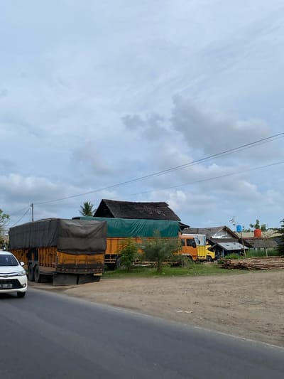 The image depicts a roadside scene in Lancang Barat, Indonesia, under an overcast sky. In the foreground, a paved road curves to the left, with a white car, license plate "B 1369 ZR," partially visible on the left edge. Beyond the road, a dirt shoulder leads to a row of parked trucks and buildings.

The most prominent subjects are two large trucks. The truck on the left is yellow and covered with a dark gray tarp, obscuring its cargo. The truck to its right is also yellow and features a bright green tarp covering its load. Further to the right, a smaller orange truck is parked, and behind it, several simple buildings with thatched or corrugated roofs are visible. In front of these buildings, a large stack of lumber lies on the ground.

The environment appears to be a semi-rural or undeveloped area, indicated by the dirt shoulder, the presence of numerous trucks, and the rustic construction of the buildings. Vegetation, including palm trees and shrubs, is scattered throughout the scene. The overall impression is one of commercial activity or transportation logistics taking place along a roadway. No people are clearly visible in the image.