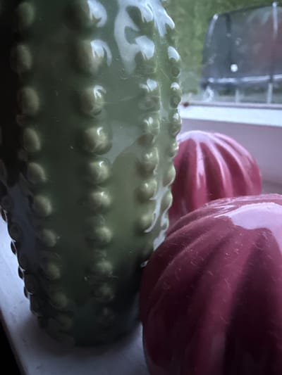 A close-up, slightly blurry, indoor shot focuses on a green, textured ceramic vase on the left, with two ridged, pink decorative objects in front of it. The background shows a blurry outdoor scene with trees and a dark structure, possibly a grill or a trampoline. The objects appear to be decorative and are arranged on a light-colored surface, possibly a windowsill. There are no people or animals visible in the image. The lighting is dim, suggesting it might be dusk or an overcast day. The scene is indoors, and the view outside the window is out of focus. There is no indication of the location in Ridabu, Norway.
