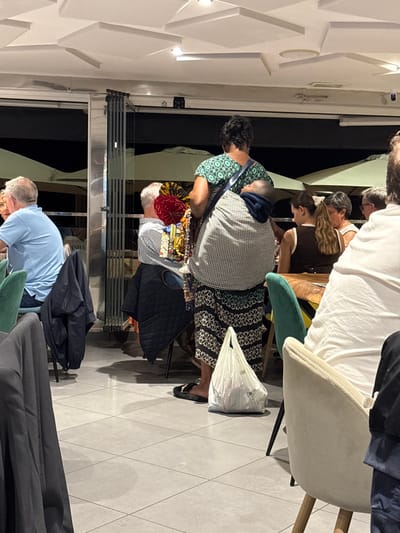 This image depicts an indoor dining area of a restaurant in Cádiz, Spain, during nighttime. The scene is dominated by a dark-skinned individual, viewed from behind, wearing a patterned teal and dark blue short-sleeved top and a long black and white patterned skirt. This individual carries a small child, whose head is visible, on their back in a grey and white checkered fabric sling. The individual also carries multiple colorful, fan-like textile items and strings of beads, possibly merchandise, in their left hand. A white plastic bag rests on the light grey tiled floor near their feet.

Several other individuals, patrons of the restaurant, are seated at tables. To the left, a fair-skinned male in a light blue polo shirt is seated, with a dark jacket draped over his chair. Another male is partially visible to his right, seated with a quilted dark jacket over his chair. Further right, a young female with light brown hair in a ponytail and an older female with short dark hair are visible from behind, seated at a wooden-topped table. A portion of a diner in a white shirt is visible in the foreground right.

The restaurant features a modern aesthetic with a light grey tiled floor, tables with dark wooden tops, and chairs upholstered in cream, green, or black. The ceiling is white with square and rectangular panels and recessed lighting fixtures. A large glass partition, appearing to be folding doors, separates the indoor dining area from an outdoor patio where light-colored umbrellas are visible against a dark background, indicating the time of day.