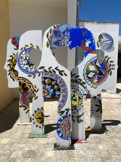 This media file captures an outdoor scene with a distinctive architectural element adorned with colorful, mosaic-like patterns. The main subject is a large, white, curved structure that appears to be part of a building or a decorative installation. It is decorated with intricate designs featuring geometric shapes, floral motifs, and abstract patterns in a variety of vibrant colors, including blue, yellow, green, purple, and orange. These decorations resemble painted tiles or mosaics, adding a lively and artistic touch to the otherwise plain white surface.
The structure is mounted on metal supports and is situated against a tiled wall that has a weathered, stone-like appearance. The scene is bathed in bright sunlight, casting distinct shadows, including the silhouette of a person standing to the right of the frame, suggesting it is daytime. The sky above is a clear, deep blue, indicating fair weather.
The overall impression is one of an outdoor public space, possibly a plaza or a walkway in a Mediterranean setting, given the tiled surfaces and bright sunlight. The artwork itself evokes a sense of cultural richness and artistic expression, possibly reflecting local craftsmanship or a public art project. The detail and vibrant colors of the painted patterns are a prominent feature, drawing the viewer's eye and suggesting a deliberate aesthetic choice for the location. There is no visible text in the image.
