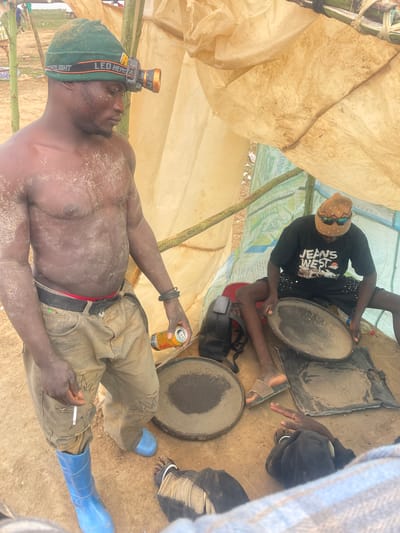 In Maiyanga, Nigeria, inside a makeshift shelter covered with tarpaulin, two men are engaged in mining-related activities. One man, lying shirtless and covered in dust, wears blue rubber boots, khaki pants, and a headlamp. He holds a beer in one hand and a cigarette in the other. The second man, seated, wears a black t-shirt and hat, operating a circular pan filled with sediment. Another similar pan sits nearby.