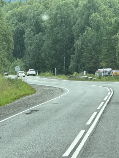 The image captures a curved asphalt road in Izvestkovyi, Russia, flanked by a dense forest of green deciduous trees. The road has a broken white line marking its center and solid white lines delineating its edges. On the right side of the road, a white trailer with the Cyrillic word "МОТОЧКА" (which can translate to "small thread" or "spool") is visible, along with a person standing near it. On the left side of the road, there are three cars: a dark-colored SUV in the distance, a white sedan, and a white SUV with its driver-side door open. A road sign with a circular white face on a gray pole is positioned between the cars. The environment appears to be a rural or semi-rural area with lush vegetation and a cloudy sky.