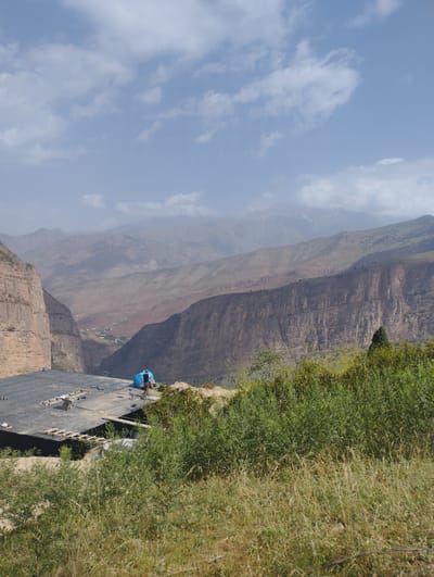 A man in dark clothing and a blue cap stands next to a large, bright blue, cylindrical object on a dark, flat surface. The surface appears to be a construction site, with wooden planks, ladders, a wheelbarrow, and ropes scattered around. The construction site overlooks a vast, rugged mountain landscape under a cloudy sky. In the distance, a small village can be seen nestled in a valley between steep, reddish-brown cliffs. The mountains stretch into the horizon, with layers of peaks fading into a hazy atmosphere. Lush green vegetation covers the foreground and edges of the cliffs, contrasting with the arid, rocky terrain. The image was taken in Chaknak, Uzbekistan.