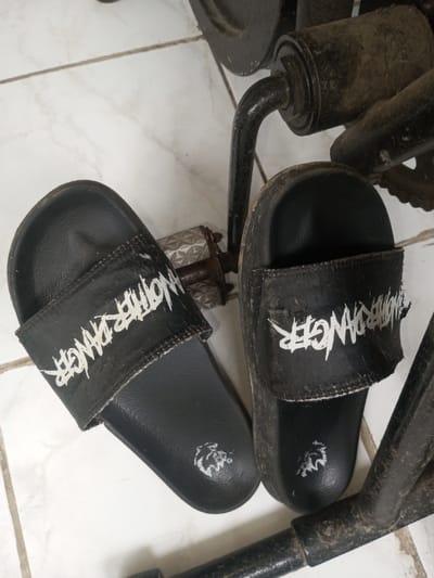 A pair of black, heavily worn slides sit near a rusty and dusty piece of exercise equipment, possibly an elliptical. The slides have "ANOTHER DANGER" printed in white on the strap and a white lion logo on the footbed. The equipment is also covered in dust and rust, particularly around the pedal area where the slides are positioned. The background is a white tiled floor with mottled patterns.