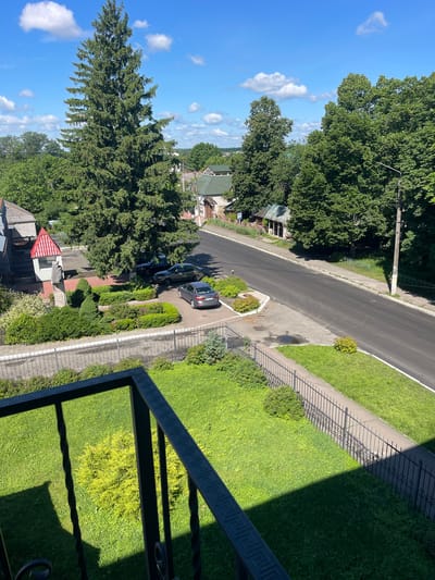 The image shows a view from a balcony overlooking a street in Makariv, Ukraine. The street is paved and appears to be moderately trafficked, with a few cars parked along the side and one driving away. There are several buildings visible, including what appears to be a small commercial establishment with a red-tiled roof and a statue in front. Trees and greenery are abundant, with a very large evergreen tree dominating the left side of the frame. The sky is blue with scattered white clouds, suggesting fair weather. The foreground is occupied by a railing and a well-maintained lawn.