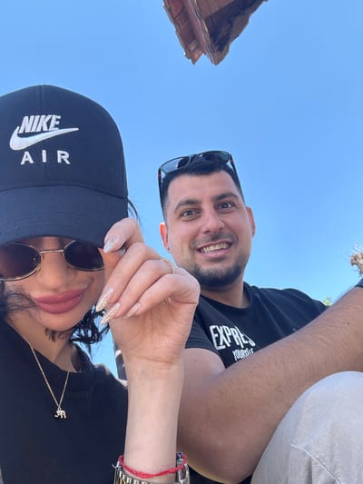 A low-angle selfie shows a man and a woman against a blue sky. A portion of a building's roof is visible in the upper left. The man, in a black T-shirt, is smiling, and his face is partially shaded by sunglasses resting on top of his head. The woman is also wearing sunglasses and a black baseball cap with "NIKE AIR" in white lettering. She has long, glittery fingernails. A gold elephant pendant hangs from her necklace. A silver wristwatch with a red string bracelet is visible on her wrist. The location is Probuda, Bulgaria, suggested by the type of architecture visible.