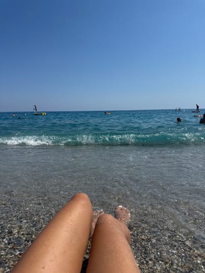 The image depicts a beach scene with a clear blue sky and turquoise water. In the foreground, a person's legs and feet are visible, resting on a pebbly shore with shallow water lapping at them. The water is transparent, revealing the pebbles beneath. In the mid-ground, the ocean stretches out with gentle waves. Several people are visible in the water, some swimming and others on paddleboards. One person is standing on a paddleboard. The overall impression is a sunny, relaxing day at the beach. The location is identified as Finale Ligure, Italy.