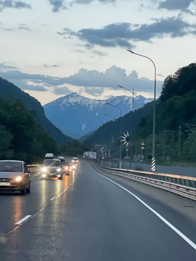 The image depicts a highway winding through a mountainous valley in Chvizhepse, Russia, during twilight or overcast conditions. The road surface is wet, reflecting the headlights of several vehicles traveling in the same direction. The vehicles include a silver sedan in the foreground on the left, and several vans and other cars further ahead, their headlights illuminated. The highway is bordered by a metal guardrail with reflective markers. Dense, dark green trees line the slopes of the mountains on either side of the valley, with the peaks of snow-capped mountains visible in the distance under a dramatic sky filled with dark, swirling clouds. Several streetlights with curved arms are positioned along the roadside. In the mid-ground, there are signs and what appear to be decorative wind sculptures. The scene suggests movement and travel along a scenic route.