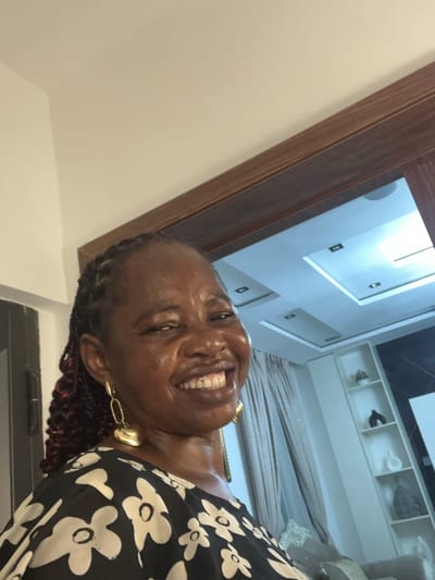 The image shows a smiling woman in a black and white floral print dress, wearing gold earrings. She is positioned in a doorway, looking into the camera. In the background, there's a room with modern lighting fixtures on the ceiling and a white shelving unit with decorative objects. A bed with greyish bedding is also visible. It is impossible to tell from the image if the location is in Mopo-Akinlade, Nigeria.