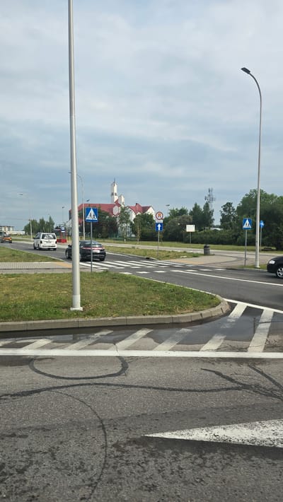 The image depicts a street scene in Suwałki, Poland, on a cloudy day. The perspective is from a vehicle, looking towards a crosswalk and a street with buildings and trees in the background.
In the foreground, a paved road with white crosswalk markings is visible. To the right, there is a curb and a grassy area. There are some dark, circular marks on the road, possibly from tire skids.
Further down the road, several cars are present, including a black car stopped at the crosswalk and a white van driving away. Traffic signs are visible along the side of the road. One sign shows a pedestrian symbol within a blue circle, indicating a pedestrian crossing. Another sign, partially obscured, appears to be a directional arrow. A sign with a red circle and a horizontal bar across it, often indicating a prohibition, is also present. In the distance, buildings with red roofs are visible amongst trees.
A tall streetlight with a curved arm stands prominently on the left side of the image. The sky is overcast with clouds. The overall impression is of a typical urban or suburban environment during daylight hours. There is no discernible text in the image.