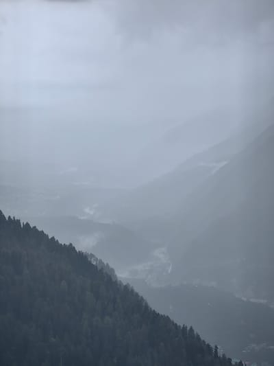 The image captures a hazy, mountainous landscape in Siumsa, India. In the foreground, a dense forest of dark green coniferous trees covers the steep slope of a mountain. The trees are densely packed, with their tops forming an uneven, jagged line against the sky.
Beyond the immediate treeline, the terrain descends into a valley. This valley is obscured by thick fog and mist, rendering distant features indistinct. A winding river or stream can be faintly discerned snaking through the valley floor, appearing as a lighter, sinuous line against the darker, mist-shrouded hills.
Further into the distance, successive layers of mountains rise, fading into the atmospheric haze. The overall color palette is muted and dominated by shades of gray, green, and blue, creating a somber and somewhat ethereal mood. The sky is overcast with heavy, uniform clouds. No people or specific structures are clearly visible due to the distance and atmospheric conditions. The scene appears to be a natural, undeveloped area, characteristic of a remote mountainous region.