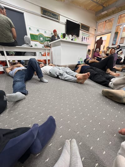 In a room in Tar-Suu, Kyrgyzstan, several people are lying down on a gray carpet. In the foreground, the legs and feet of several individuals are visible, some wearing socks of various colors and textures. In the midground, two men are lying on their backs on the carpet. The man on the left wears a blue t-shirt and jeans with gray socks. The man on the right wears a white t-shirt and gray sweatpants. Further back, other individuals are seated or lying down, some with heads covered. A woman wearing a red headscarf is visible standing at a white counter. Behind the counter, a banner with a green logo and Arabic text is displayed. The room has large windows and an interior wall with framed calligraphy and informational posters.