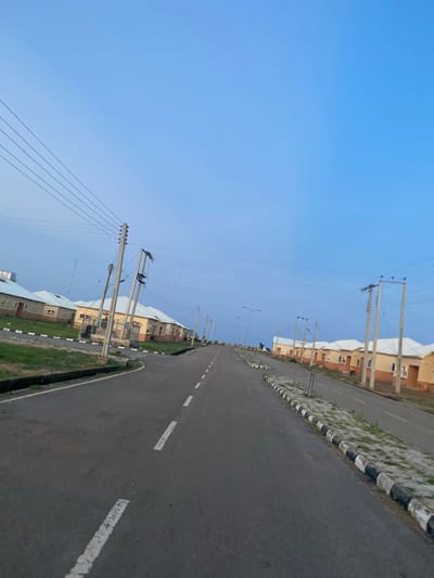 The media file displays an outdoor scene of a street in Dumnga, Nigeria, under a clear blue sky, likely during the daytime. The perspective is from a slightly elevated angle, looking down a paved road. The road features white dashed lines and a solid white line marking the lane. Along the left side of the road is a raised median with alternating black and white concrete borders, with sparse green vegetation growing through it.
On the right side of the road, there are several buildings with a light orange-brown facade, featuring rectangular windows. These appear to be residential or commercial structures arranged linearly. Parallel to the road and stretching into the distance are numerous utility poles carrying power lines. The power lines are taut and extend across the sky.
There are no people visible in the image, and the overall scene suggests a quiet, possibly newly developed or maintained area. The weather appears to be fair and clear, with no clouds present. The time of day is likely afternoon or late morning, given the brightness of the sky. No text is discernible in the image.