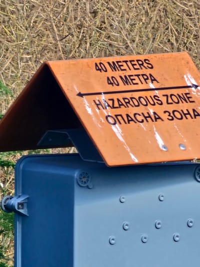 An orange sign with black lettering is affixed to the top of a grey utility box. The sign reads: "40 METERS 40 METPA HAZARDOUS ZONE ОПАСНА ЗОНА". An arrow points to the left. The grey utility box has several circular indentations on its surface and a bracket with a screw on its left side. The background is a blur of dry grass and foliage. The sign indicates a hazardous zone extending 40 meters from this point. This scene is located in Kolarovo, Bulgaria.