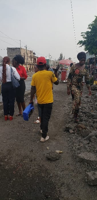 This image depicts an outdoor street scene in Buzove, Democratic Republic of the Congo, under an overcast sky. The ground is unpaved, consisting of dark grey dirt, gravel, and loose rocks, indicating an uneven and rugged surface.

In the midground, a male subject is viewed from behind, wearing a yellow t-shirt, dark trousers, and a red baseball cap. He carries a blue plastic container in his left hand and an indistinct white object in his raised right hand, walking towards the right side of the frame.

To the right, a female subject walks towards the left, wearing a long, dark patterned dress with circular motifs and a dark headwrap, navigating the uneven ground.

On the left side of the frame, a group of individuals is present. One female wears a white long-sleeved shirt, dark skirt, and red high-heeled shoes, carrying a light-colored shoulder bag. Another female is partially visible behind her, wearing a red short-sleeved top and a dark skirt. Another individual's lower legs and feet, wearing white sandals, are visible behind the male subject in yellow.

In the background on the left, multiple-story buildings constructed of concrete blocks are visible. On the right, a small tree and a red umbrella, possibly part of a market stall, are present. Utility lines are visible against the sky.