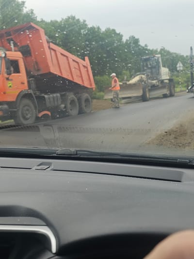 In Solnechnoe, Russia, an orange dump truck is unloading material onto a paved road. A road grader is positioned behind the dump truck, and a worker in an orange vest is standing near it. The road appears to be under construction, with loose gravel and dirt present. Trees line the background, and the sky is overcast. The photo is taken from inside a vehicle, with the windshield partially visible in the foreground.