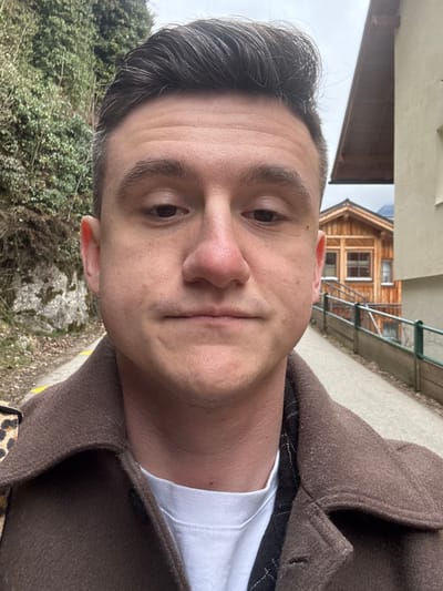 A male subject is depicted in a close-up, head-and-shoulders selfie, positioned on a pathway within the town of Hallstatt, Austria. The subject, an adult male with short dark hair, dark eyebrows, and a light complexion, maintains a neutral facial expression. He wears a brown collared coat over a white crew-neck shirt, with a dark, possibly quilted, garment visible beneath the coat collar. A leopard print pattern is discernible on his left shoulder, indicative of an accessory strap or detail.

The background reveals a narrow, light-colored pathway, possibly paved or gravel, extending behind the subject, with faint yellow markings along its edge. To the subject's left, a steep, natural rock wall densely covered with green vegetation rises. To the subject's right, a partial view of a light-colored building with a dark roof is visible. Further along and to the right, at a lower elevation relative to the pathway, stands a traditional multi-story wooden chalet-style building with multiple windows, featuring natural wood siding. A wooden fence or railing separates the pathway from this lower structure. The sky is overcast, providing uniform, diffused lighting to the scene.