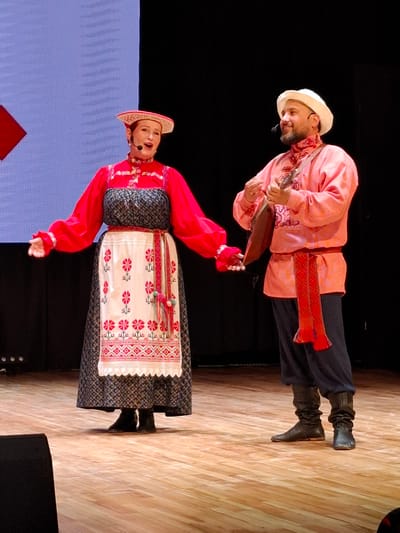 In a dimly lit theater, two performers, a woman and a man, stand on a wooden stage in front of a dark backdrop and a large screen displaying a light blue and red geometric pattern. The woman, on the left, wears a traditional red and white embroidered Russian folk costume. Her costume includes a red blouse with voluminous sleeves, a dark patterned skirt worn over a white apron adorned with red floral embroidery and intricate geometric patterns, and a tall, ornate headdress. She holds her arms out to the sides and has a microphone near her mouth, suggesting she is singing or speaking. The man, on the right, is dressed in a vibrant pink embroidered shirt, dark pants, and black boots. He wears a white cap and has a beard. He is playing a traditional stringed instrument, possibly a balalaika, and also has a microphone. The stage lighting illuminates the performers and the wooden floor. The scene likely depicts a cultural performance or concert. While the location is mentioned as Kungur, Russia, there are no specific visual cues in the image to confirm this detail.