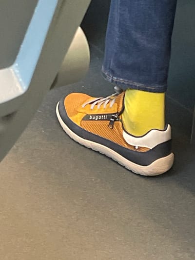 A close-up, ground-level shot shows a person's foot wearing a yellow mesh sneaker with white laces, a dark blue sole, and a white heel accent. The sneaker has a zipper with a "bugatti" logo. The person is wearing dark blue jeans and bright yellow socks. The foot is resting on a dark gray, textured floor. In the background, a portion of a light gray bench or seat is visible. The setting appears to be indoors, possibly in a public transportation hub, given the seating and flooring. The photograph was taken in Lahošť, Czechia.